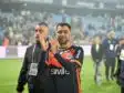 Male athlete in an orange-and-black jersey applauds on a football pitch with a cheering crowd in the blurred stadium behind him.