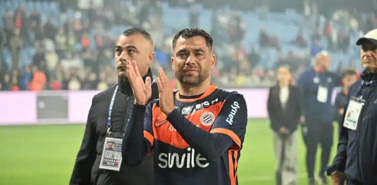 Male athlete in an orange-and-black jersey applauds on a football pitch with a cheering crowd in the blurred stadium behind him.