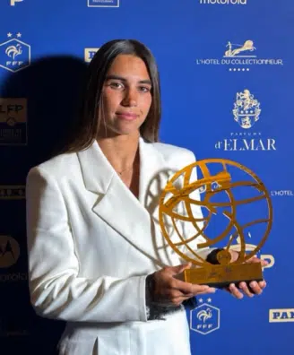 Woman in a white blazer holding a gold globe-like award at a blue sponsor backdrop.