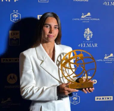 Woman in a white blazer holding a gold globe-like award at a blue sponsor backdrop.