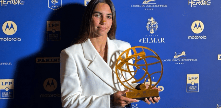 Woman in a white blazer holding a gold globe-like award at a blue sponsor backdrop.