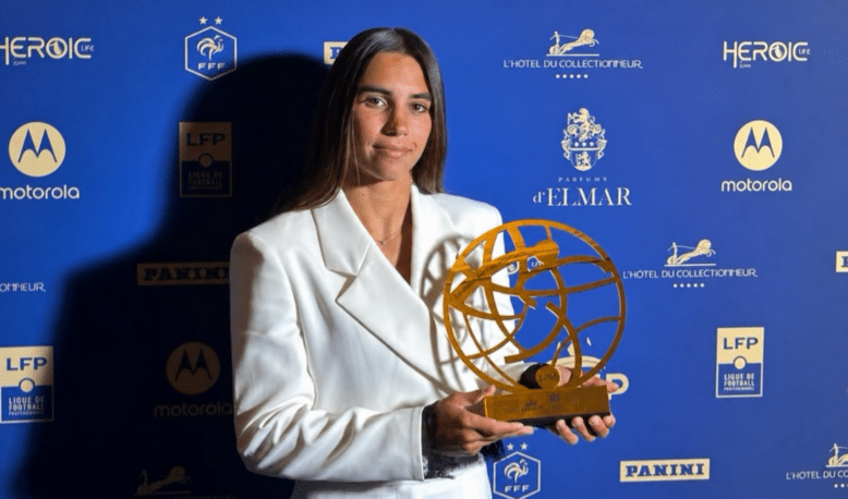 Woman in a white blazer holding a gold globe-like award at a blue sponsor backdrop.