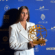 Woman in a white blazer holding a gold globe-like award at a blue sponsor backdrop.
