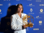 Woman in a white blazer holding a gold globe-like award at a blue sponsor backdrop.