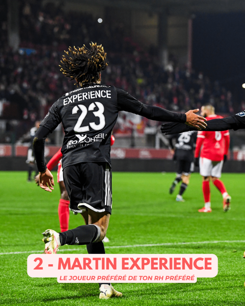 Soccer player with dreadlocks, wearing a black jersey numbered 23 reading 'EXPERIENCE', runs on a bright green field with a crowded stadium in the background; a bottom banner reads '2 - MARTIN EXPERIENCE'.