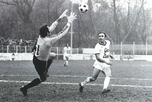 Goalkeeper dives to reach the ball as an attacker advances during a black-and-white soccer match.