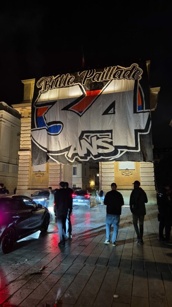 Night scene of a large banner draped over a stone archway, with people standing and cars on a wet street at night.