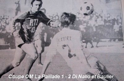 Soccer match moment: a dark‑kit attacker challenges a white‑kit goalkeeper for the ball as a crowd watches; caption reads 'Coupe OM La Paillade 1 - 2 Di Nallo et Baulier'.