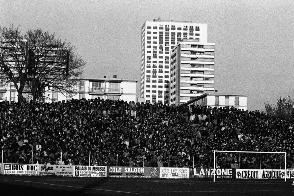 Crowd of spectators packed into a stadium stands, banners along the front, with high-rise buildings in the background.