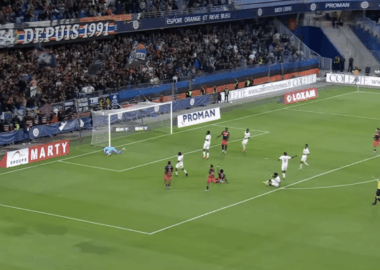 Soccer match; goalkeeper dives to stop a shot near the goal as attackers and defenders crowd the area.