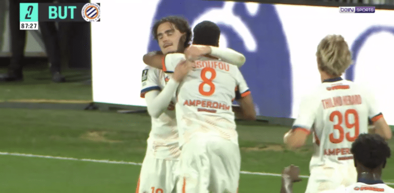 Two soccer players in white kits hug and celebrate on the green pitch after a goal.