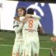 Two soccer players in white kits hug and celebrate on the green pitch after a goal.