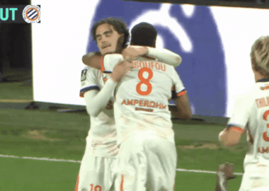 Two soccer players in white kits hug and celebrate on the green pitch after a goal.