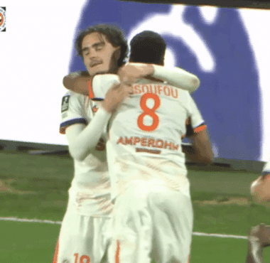 Two soccer players in white kits hug and celebrate on the green pitch after a goal.