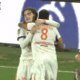 Two soccer players in white kits hug and celebrate on the green pitch after a goal.