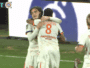 Two soccer players in white kits hug and celebrate on the green pitch after a goal.
