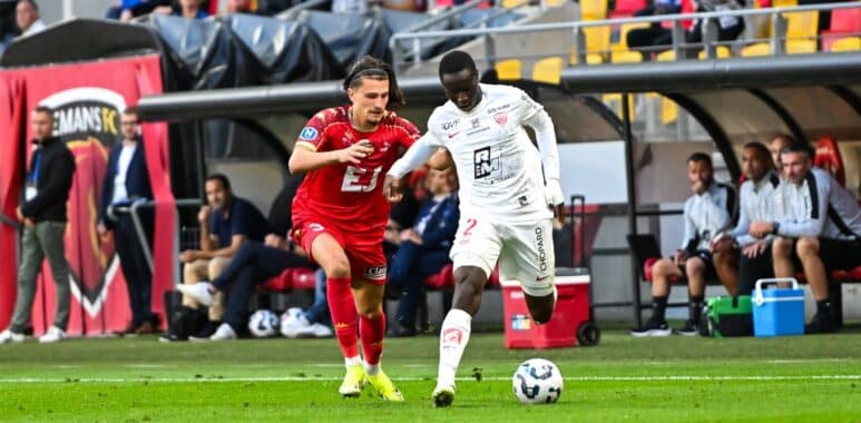 Two soccer players&mdash;one in red, one in white&mdash;racing for the ball on a green pitch during a match, with teammates and fans in the background of the stands and dugout.