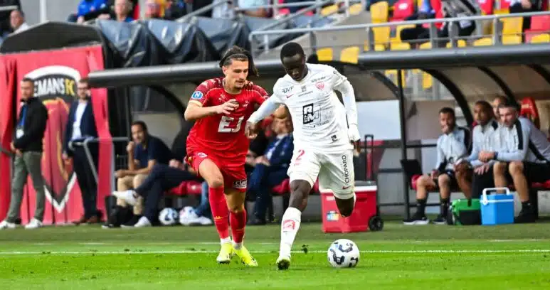 Two soccer players&mdash;one in red, one in white&mdash;racing for the ball on a green pitch during a match, with teammates and fans in the background of the stands and dugout.
