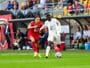 Two soccer players&mdash;one in red, one in white&mdash;racing for the ball on a green pitch during a match, with teammates and fans in the background of the stands and dugout.