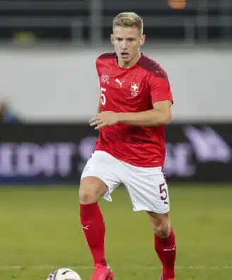 Soccer player in a red jersey with number 5 dribbling the ball on a grass field; Swiss team crest visible on chest.