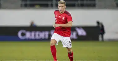Soccer player in a red jersey with number 5 dribbling the ball on a grass field; Swiss team crest visible on chest.