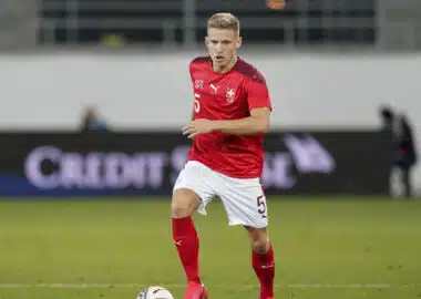 Soccer player in a red jersey with number 5 dribbling the ball on a grass field; Swiss team crest visible on chest.