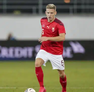 Soccer player in a red jersey with number 5 dribbling the ball on a grass field; Swiss team crest visible on chest.