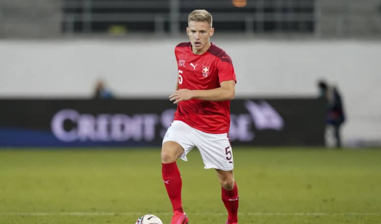 Soccer player in a red jersey with number 5 dribbling the ball on a grass field; Swiss team crest visible on chest.