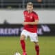 Soccer player in a red jersey with number 5 dribbling the ball on a grass field; Swiss team crest visible on chest.