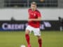 Soccer player in a red jersey with number 5 dribbling the ball on a grass field; Swiss team crest visible on chest.