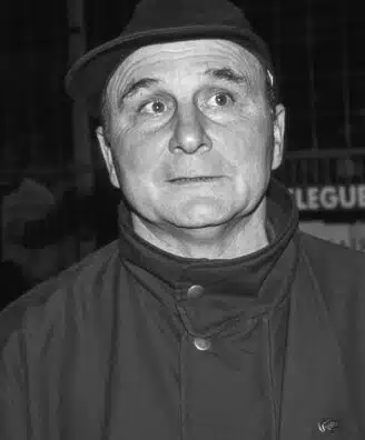 Black-and-white photo of a man in a fedora and high-collar coat, eyes wide in a startled expression.