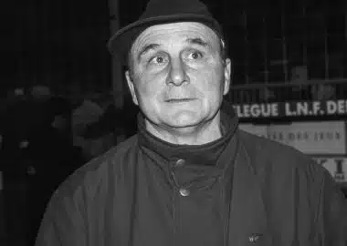 Black-and-white photo of a man in a fedora and high-collar coat, eyes wide in a startled expression.