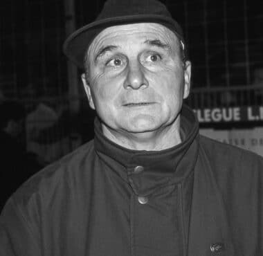 Black-and-white photo of a man in a fedora and high-collar coat, eyes wide in a startled expression.