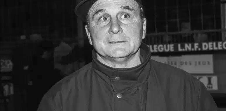 Black-and-white photo of a man in a fedora and high-collar coat, eyes wide in a startled expression.