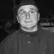 Black-and-white photo of a man in a fedora and high-collar coat, eyes wide in a startled expression.