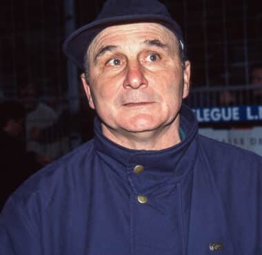 Man in a blue uniform/jacket and dark cap, looking to the side with a surprised expression at night outdoors.