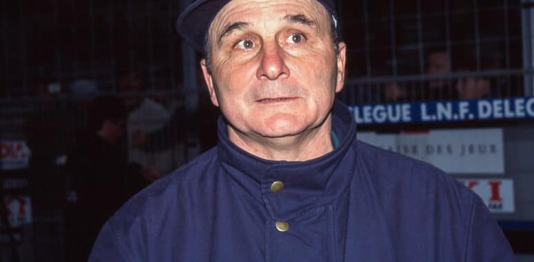 Man in a blue uniform/jacket and dark cap, looking to the side with a surprised expression at night outdoors.