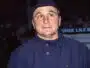 Man in a blue uniform/jacket and dark cap, looking to the side with a surprised expression at night outdoors.