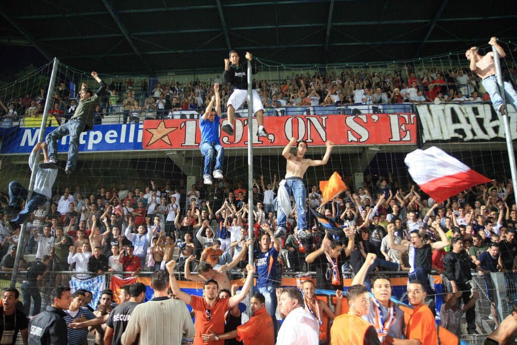 Crowd of soccer fans in a stadium behind protective netting, cheering and waving banners as security guards stand in front.] }
