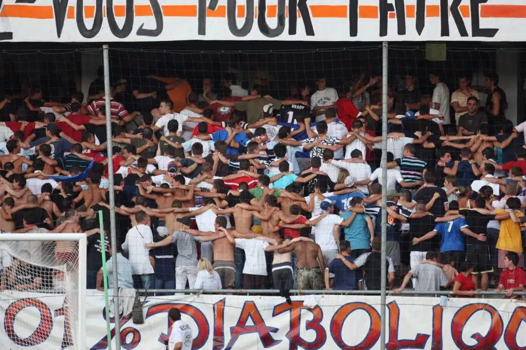 Crowd of football fans with arms linked, standing behind a goal net in a stadium, with a large banner above.