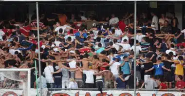 Crowd of football fans with arms linked, standing behind a goal net in a stadium, with a large banner above.
