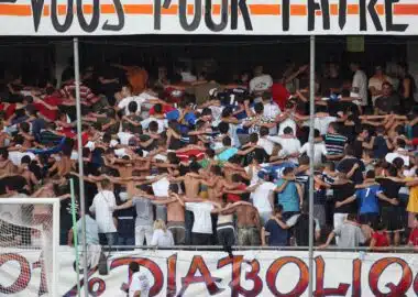 Crowd of football fans with arms linked, standing behind a goal net in a stadium, with a large banner above.