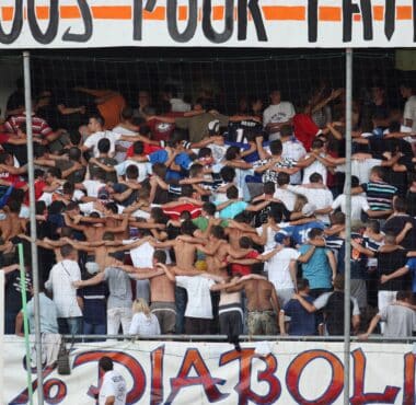 Crowd of football fans with arms linked, standing behind a goal net in a stadium, with a large banner above.