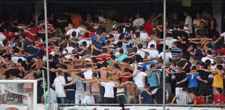 Crowd of football fans with arms linked, standing behind a goal net in a stadium, with a large banner above.