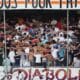 Crowd of football fans with arms linked, standing behind a goal net in a stadium, with a large banner above.