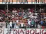 Crowd of football fans with arms linked, standing behind a goal net in a stadium, with a large banner above.