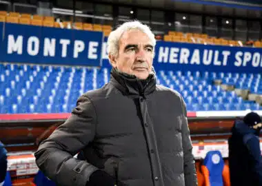Older man in a gray winter coat stands in a sports stadium with blue seats and large white lettering behind him.