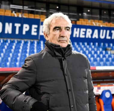 Older man in a gray winter coat stands in a sports stadium with blue seats and large white lettering behind him.
