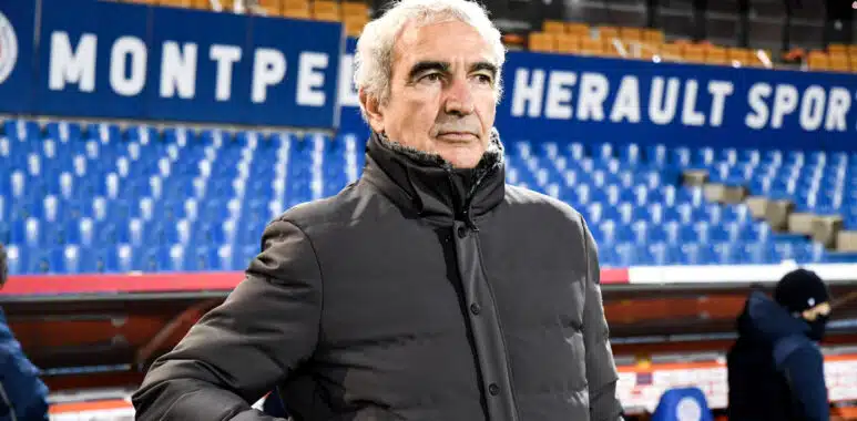 Older man in a gray winter coat stands in a sports stadium with blue seats and large white lettering behind him.