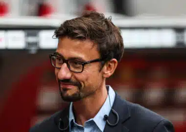 Man with dark hair and glasses, wearing a blue shirt and dark blazer, looking to the left outdoors.